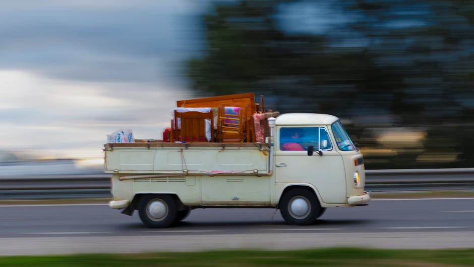 A vintage cream-colored van with a flatbed cargo area is shown in motion on a road, captured during the loading process for a home relocation. The flatbed holds various household furniture pieces including wooden chairs, a small table, and wrapped packages, secured with plastic wrap and packing materials. Several cardboard boxes and additional wrapped items are also visible, indicating packing for a move. The van is positioned on the right side of the image, with the background showing a blurred landscape of trees and cloudy sky, suggesting the vehicle is moving at speed. The scene depicts a typical scene of furniture transport during a moving service, with the van parked close to a property entrance or loading area. This image exemplifies the logistics involved in house removals, such as loading household belongings onto a van for relocation, as coordinated by companies like Man with Van Paddington, especially for moves within the Sussex Gardens area. The overall lighting indicates daytime, emphasizing the practical aspects of packing and transport in a professional moving context.