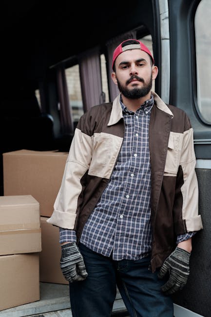 A young man wearing a beige knitted beanie, a green padded vest over a dark blue jacket with white stripes on the sleeves, and black gloves, is seated on the edge of an open moving van. Inside the van, several cardboard boxes of various sizes are arranged on the floor and against the sides, some wrapped in plastic for protection. The interior of the van appears spacious with a dark ceiling and walls, and the background suggests an urban street environment. The man appears to be in the process of a house relocation, with the boxes ready for loading or unloading, reflecting the packaging and moving process. This scene is associated with home removals and furniture transport services provided by Man with Van Paddington, highlighting the logistical aspects of packing and loading during a move involving small-van strategies in Sussex Gardens.