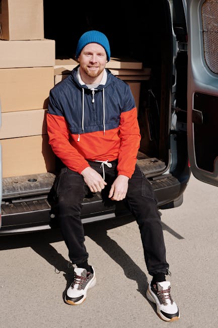 A man wearing a beige knit beanie, a green and navy blue padded jacket, and black gloves stands beside a black moving van during a home relocation. Inside the van, cardboard boxes are visible, includi
