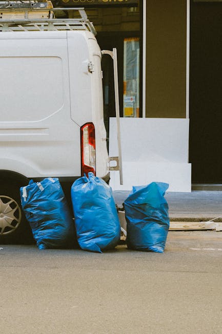 A white commercial van parked on a city street during daytime, with its sliding side door partially open, revealing a glimpse of the vehicle's interior. Three large blue plastic bags filled with waste are positioned on the pavement directly adjacent to the rear of the van, suggesting disposal of household or packing materials related to home relocation or furniture transport. The bags are situated on the street surface, which appears clean, and are likely awaiting collection or disposal as part of the moving process. Behind the van, there is a building with a glass door and window, indicating an urban environment. The scene is well lit with natural daylight, emphasizing the practical aspects of loading and waste management during house removals or relocation services, as handled by companies like Man with Van Paddington.