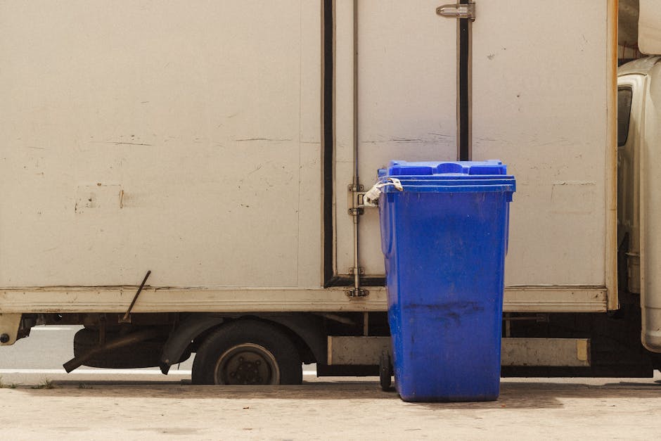 A white commercial van parked on a city street during daytime, with its sliding side door partially open, revealing a glimpse of the vehicle's interior. Three large blue plastic bags filled with waste are positioned on the pavement directly adjacent to the rear of the van, suggesting disposal of household or packing materials related to home relocation or furniture transport. The bags are situated on the street surface, which appears clean, and are likely awaiting collection or disposal as part of the moving process. Behind the van, there is a building with a glass door and window, indicating an urban environment. The scene is well lit with natural daylight, emphasizing the practical aspects of loading and waste management during house removals or relocation services, as handled by companies like Man with Van Paddington.
