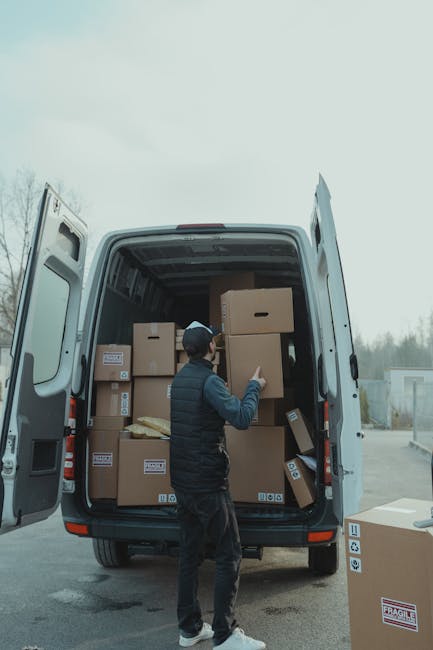 A young man with a beard and wearing a red cap backwards, a brown and beige jacket over a blue checkered shirt, and grey striped gloves, is sitting on the back of a van with an open tailgate during a 