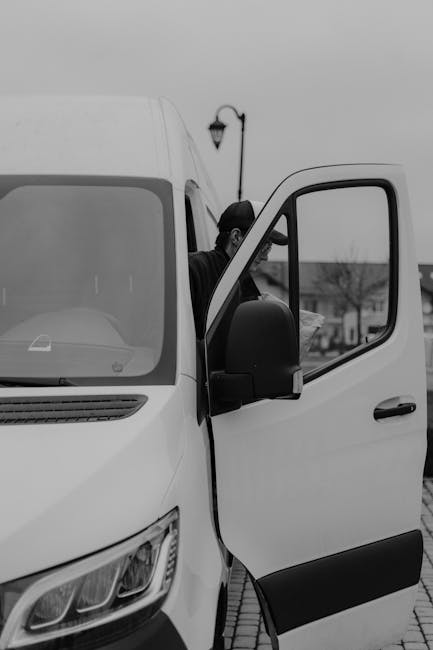 A black-haired man wearing dark clothing is seen loading a large, white moving van with the driver's side door open, revealing the interior. The van is parked on a paved street or driveway in a residential area, with a vintage-style street lamp and some trees visible in the background. Inside the van, various packing materials such as cardboard boxes, wrapped furniture, and plastic coverings are visible, indicating a recent or ongoing home relocation process. The scene is lit by natural daylight, capturing the practical aspects of furniture transport and packing for house removals, with the focus on the loading process handled by the staff from Man with Van Paddington as part of their same-day removal services.