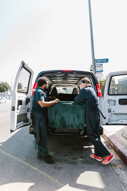 Two men wearing dark uniforms with utility belts are loading a large, dark green upholstered piece of furniture into the back of a black van parked on a street in Paddington. The van's rear doors are open, revealing an interior lined with black framework and a padded surface. One man is holding the furniture steady while the other is pushing it inside. The scene is set outdoors under bright daylight, with a clear sky, a street sign reading ‘Somerset’ visible in the background, and a curb with a pink-painted section along the pavement. The image captures the process of furniture transport during a home relocation, with packing and moving equipment in use, supported by a professional removal service such as Man with Van Paddington, highlighting the loading process in a typical urban environment.