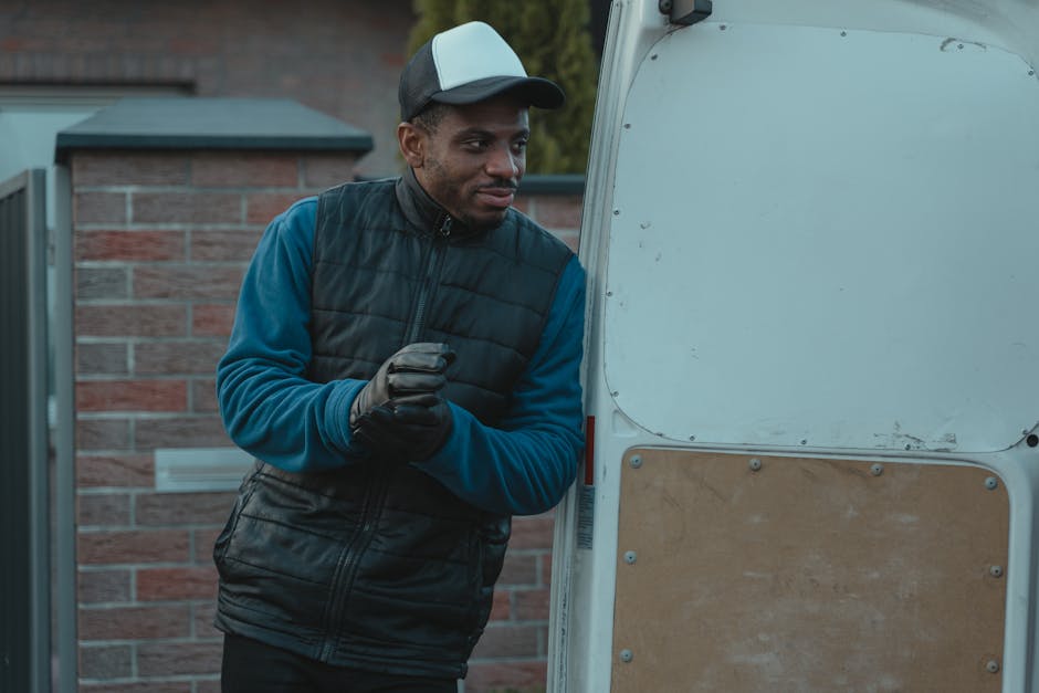 A man dressed in a blue long-sleeve shirt, a black padded vest, and a black and white cap stands outdoors next to a white van with its back doors open. He is wearing black gloves and appears to be in the process of loading or unloading; he is holding his hands together and looking towards the open van. Inside the van, partially visible, are cardboard boxes, and wrapping materials are visible around the entryway. The background shows a brick building and some greenery, with natural daylight illuminating the scene, indicative of typical house removal logistics and packing processes, supporting professional furniture transport and home relocation services offered by Man with Van Paddington.