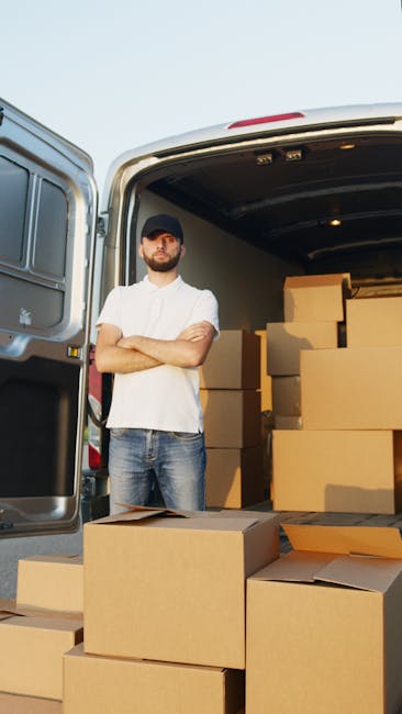 A man with a beard wearing a dark baseball cap, white t-shirt, and jeans stands with arms crossed in front of an open van door, which is filled with neatly stacked cardboard boxes for a home relocation or removals service. The boxes vary in size, with some placed inside the van and others on the ground nearby, ready for loading or unloading. The van's interior is visible with a few more boxes and packing materials, such as plastic wraps or packing paper. The scene takes place outdoors, under natural daylight, on a paved surface. This image relates to the process of furniture transport, packing, and loading involved in professional removals, as handled by Man with Van Paddington, supporting urgent eviction moveouts and house clearance services listed on the website https://manwithvanpaddington.com for efficient, legal moving logistics.