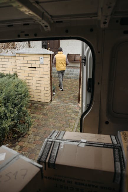 A man with a beard wearing a dark baseball cap, white t-shirt, and jeans stands with arms crossed in front of an open van door, which is filled with neatly stacked cardboard boxes for a home relocation or removals service. The boxes vary in size, with some placed inside the van and others on the ground nearby, ready for loading or unloading. The van's interior is visible with a few more boxes and packing materials, such as plastic wraps or packing paper. The scene takes place outdoors, under natural daylight, on a paved surface. This image relates to the process of furniture transport, packing, and loading involved in professional removals, as handled by Man with Van Paddington, supporting urgent eviction moveouts and house clearance services listed on the website https://manwithvanpaddington.com for efficient, legal moving logistics.