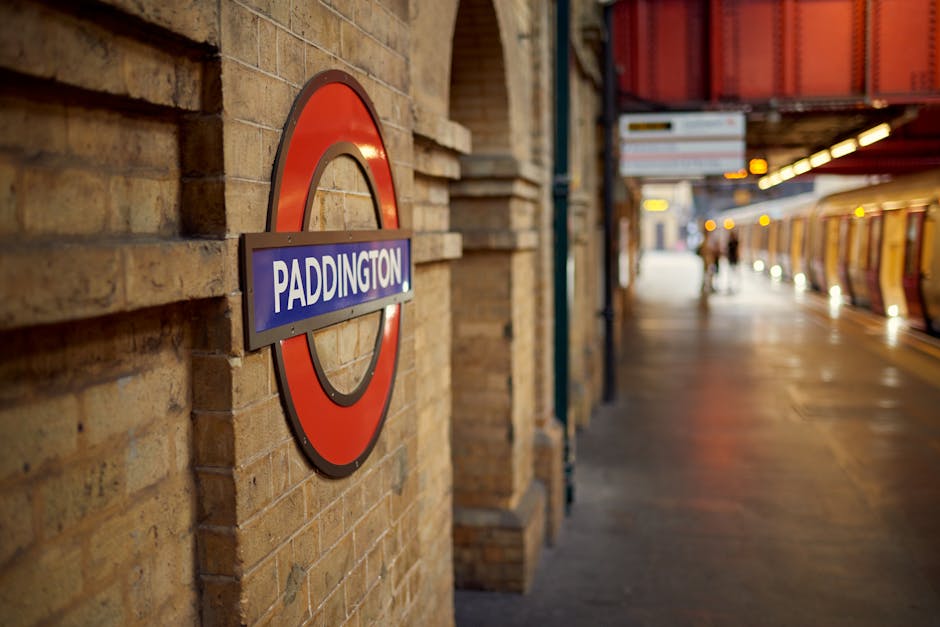 Close-up of a municipal London Underground roundel sign with a blue rectangular strip displaying the station name 'Paddington' in white letters, mounted on a brick wall of a station platform. In the background, the platform extends with a few blurred figures of passengers waiting near the edge, and a train with yellow and orange exterior is stationary along the platform. The scene is lit by overhead lighting, creating a warm, ambient atmosphere indicative of an early evening setting. The platform appears clean and organized, with safety markings on the ground, and the environment suggests a typical urban London station engaged in daily passenger transport and home relocation activities. Man with Van Paddington occasionally assists with furniture transport and packing during these stations involving comprehensive moving processes.