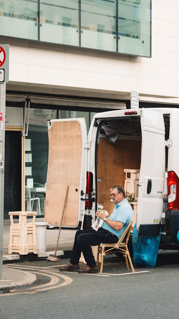 A man wearing glasses and a light blue shirt is seated on a wooden chair beside an open white moving van parked on a city street in Paddington. The van's cargo area contains large cardboard boxes and wooden furniture, with some items wrapped in protective materials such as plastic wrap and fabric blankets. A partially lifted wooden panel is propped against the back of the van, suggesting ongoing packing or furniture loading. The scene shows the man reading a document or instructions, likely related to a home relocation, as part of a furniture transport and packing process. In the background, a modern building with glass windows and a concrete facade is visible, along with a street sign on a pole nearby. The overall setting indicates a professional removals operation by Man with Van Paddington, involving careful handling of household items during a house move or furniture transport within an urban environment.